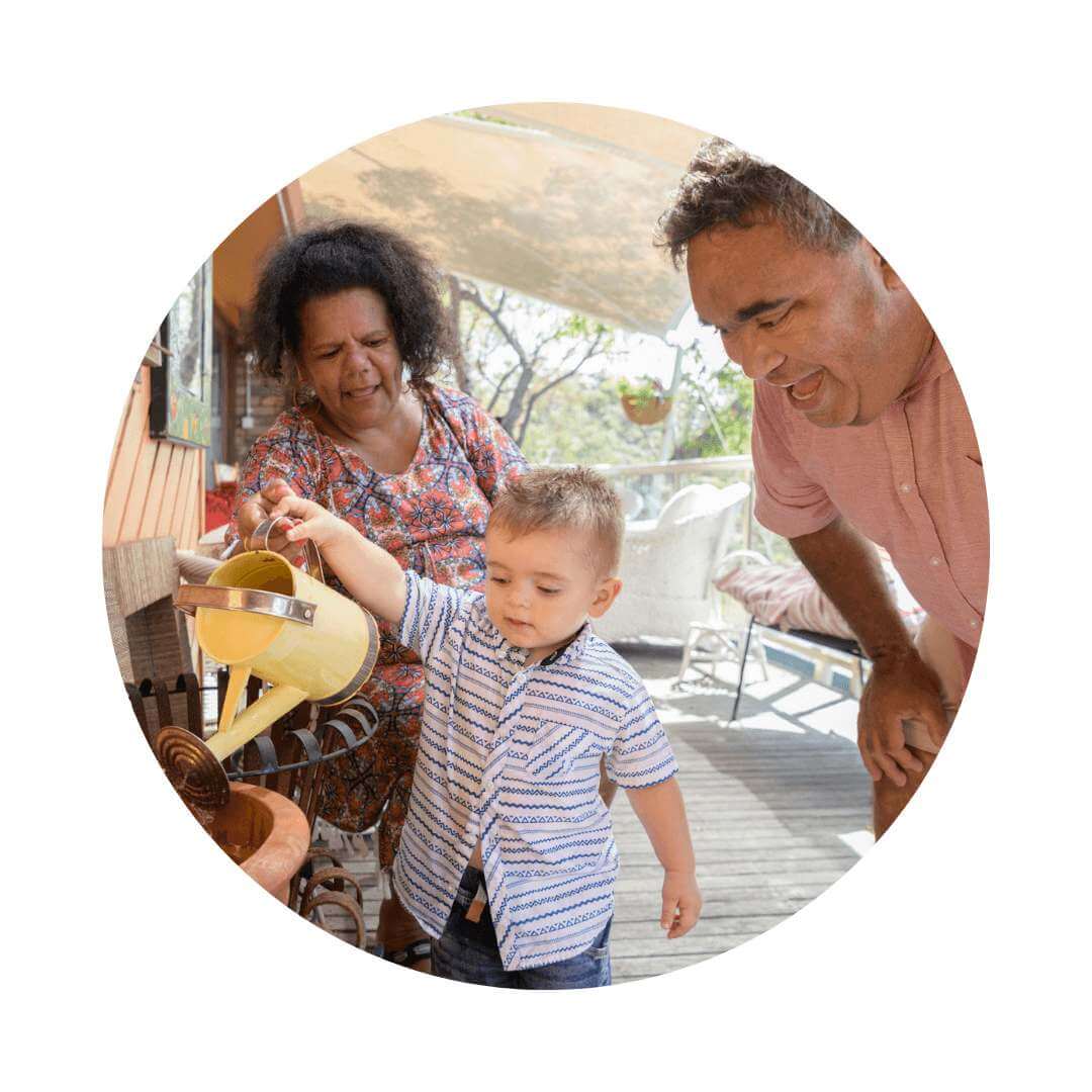 Young child in blue shirt watering a pot with a female and male watching with encouragement