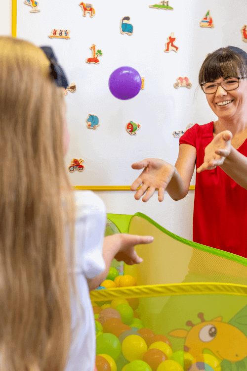 A woman in a red top throws a small purple ball to a young girl